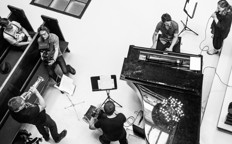 Et sort-hvid billede med et flygel placeret lidt til højre for midten med jazzmusikere omkring. Koncerten foregår på kirkegulvet i Eliaskirken på Vesterbro, København.