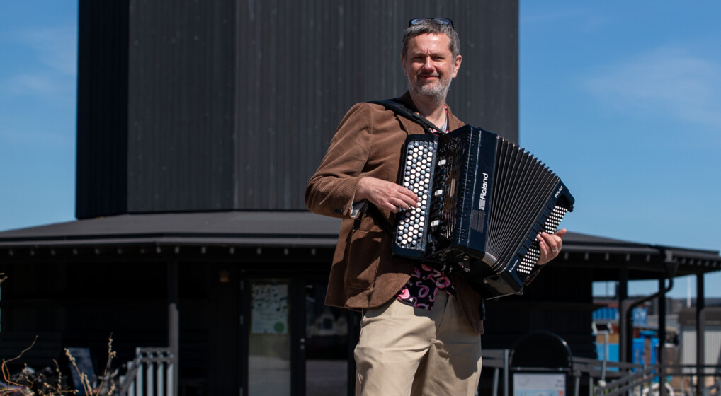 A photo of musician Martin Bennedbo in a brown corderoy jacket  with an accordion in his hands in front of Tiny Church, Nordhavn