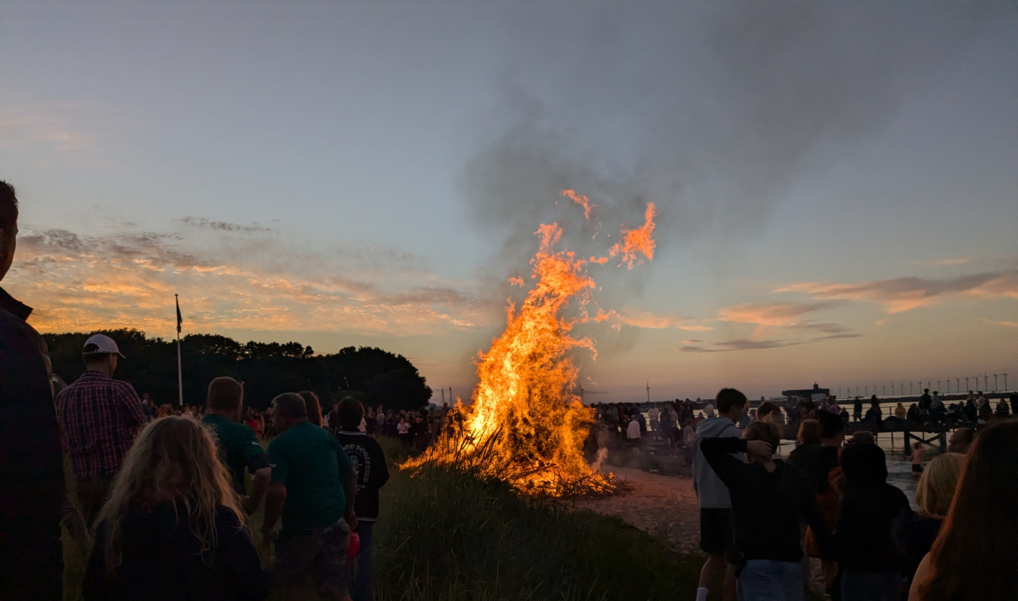 Et sankthansbål buldrer opad mod den lyse nattehimmel med mennesker forsamlet omkring