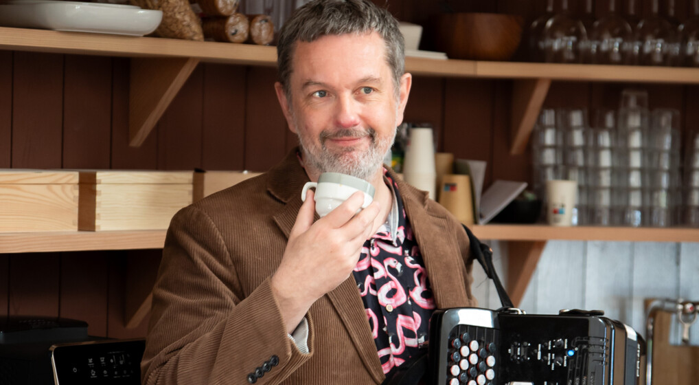 A photo of musician Martin Bennedbo in a brown corderoy jacket with a coffee cup in one hand and an accordion in the other