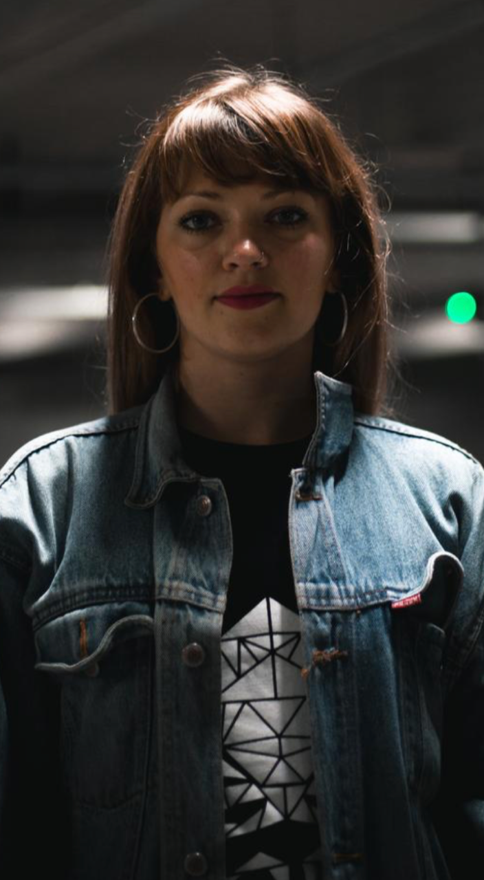 Anastasija Olescuka er fotograferet i en parkeringskælder under et hvidt lys. Hun har en denimjakke på og kigger direkte ind i kameraet