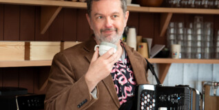 A photo of musician Martin Bennedbo in a brown corderoy jacket with a coffee cup in one hand and an accordion in the other