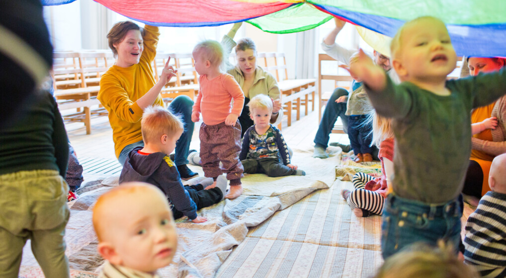 Babyrytmik En flok børn og et par voksne leger under en faldskærm i flotte farver