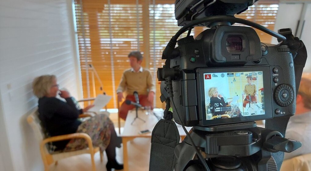 I forgrunden kamera i baggrunden to mennesker i en interviewsituation
