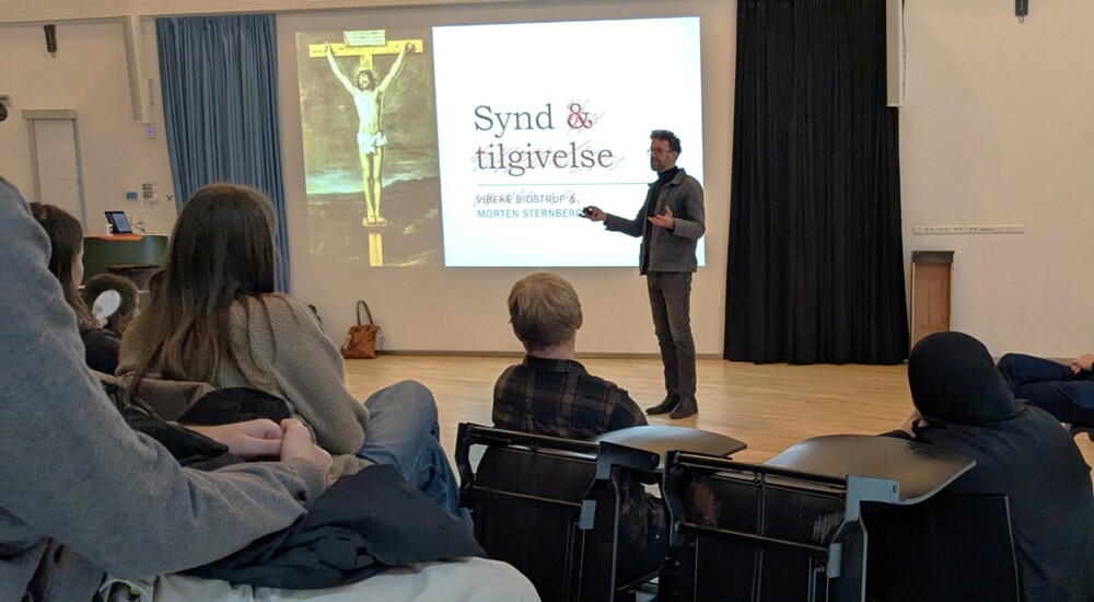 Morten sternberg, sognepræst, står foran en lærred hvor hans foredrag bliver præsenteret. I forgrunden sidder gymnasieelever med ryggen til kameraet og lytter interesseret.