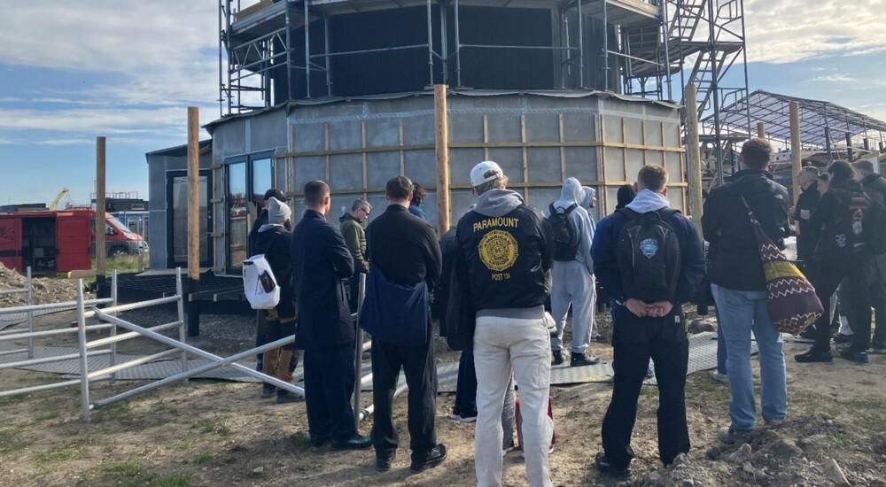 Tømrerelever står udenfor. Foran en 12-kantent bygning under færdiggørelse med stillads.