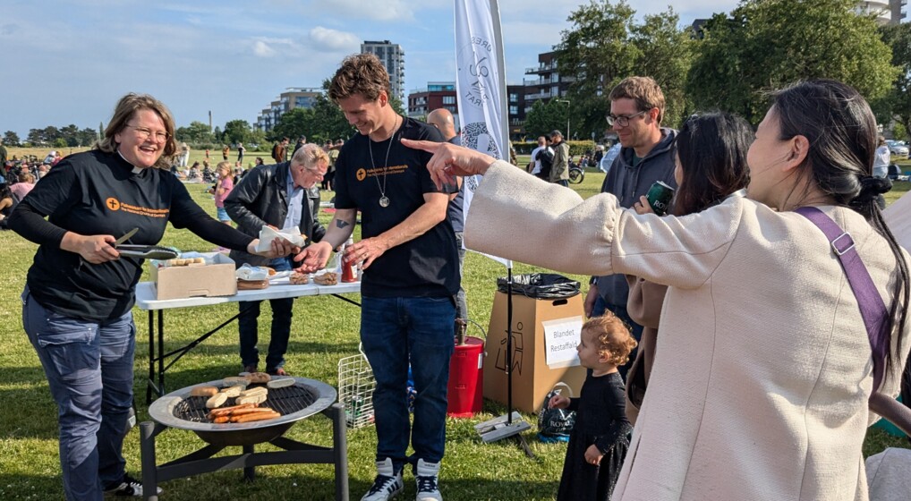 To personer fra Folkekirken for Internationals står ved en grill på Amager Strand og griner og samtaler med deltagere ved et midsummer-arrangement